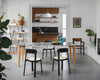 A beautiful and bright dining room. Dining table and dining chairs. Two black dining chairs with light seats in the foreground. Behind the dining table are another two dining chairs. Light concrete floor. A opening into a kitchen in the background.
