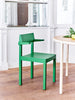 Interior image. Narrow shot. A green dining chair with armrests. Next to it we see a small part of a dining table in the colour ivory. Wooden floors. White walls in the background.