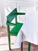 Interior in situ shot. Green dining chair with arm rests next to a table with table cloth on top. Narrow shot. Wooden floor. White wall in the background.