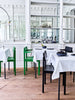 Interior shot from a bright and beautiful restaurant. Green and black dining chairs next to tables with tablecloth on top. Wooden floor. White wall with big windows in the background.
