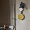 Interior shot. A wall lamp hanging on a beige coloured wall. The lamp has a few disc shaped forms. The colours of the discs are blue, black, steel and gold.