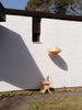 Exterior shot. A pendant light and a stool in front of a white house. Sunny day. The light is made from bendt pine veneer and the stool is made from solid pine. Some shadow play.