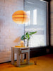 Narrow shot from inside. A big window and a white brick wall in the background. Orange carpet. On the floor in front of the wall stands the 003 Stillts side table in its boxy and adorable style. Above it hangs the ball shaped 1002 Hans pendant light. It is made from pine venner and the light is switched on.