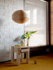 Photo shot inside. Narrow shot. On the floor next to a brick wall and window is the 003 Stillts side table. Some small home items are on its top. Above the sidetable hangs the 1002 Hans Pendant. It is shaped as a ball and made from pine veneer.