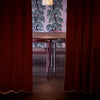 Interior photography. Dining room. Misterious atmosphere. Long burgundy curtains on the left and right. Behind the curtains is a dining table and two dining chairs. A wall with a flower print wallpaper in the background.