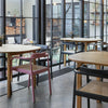 Bright and cosy cafeteria. Three small tables in light wood. Around the tables are elegant dining chairs with armrests. Chairs are in pink, black and dark blue. Gray concrete floors. Very large windows in the background.
