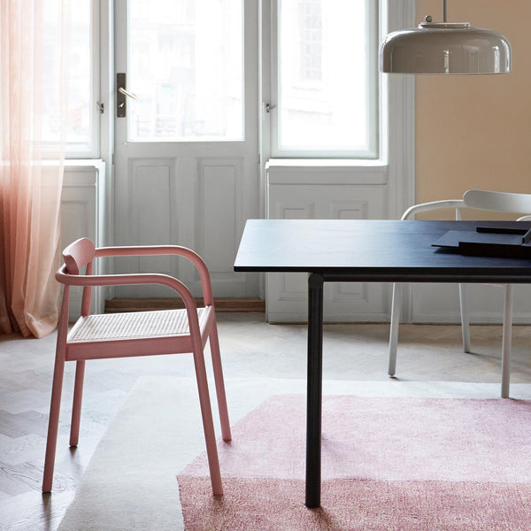 Bright dining room. Beautiful furniture. Pink dining chair with armrests. Light pink rug. Dark dining table. Light grey dining room light above table. White windows and door in the background.