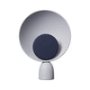 Product photo. A cute circular table lamp on a white background. The table lamp is in light grey colour. The light source is in the middle of the lamp and also circular. Light source is in navy blue colour.