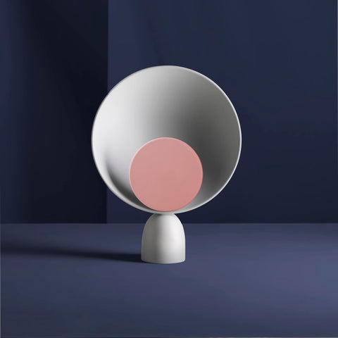 A sculptural table lamp. The lamp is in light grey colour. The light diffuser is circular and in the middle. The diffuser is in light pink colour. The background is navy blue.