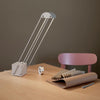 Narrow Insitu shot. On a corner of a light wooden table top sits a light grey, modern table lamp. A backrest of a pink dining chair visible behind the table.