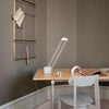 Interior image. Light grey dining chair in front of a dining table. Dining table has a wooden table top and metal feet. A white modern desk lamp on the table along with various small objects. Grey walls. A grey and brass pinboard on the wall.