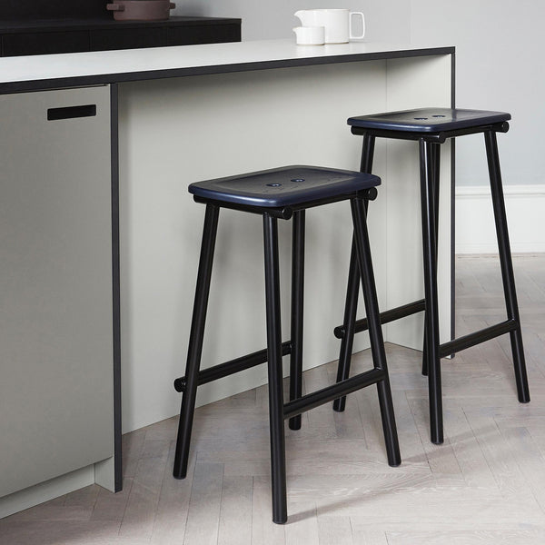 Mynd innan úr eldhúsi. Tveir dökkir kollar í sama stíl standa hlið við hlið fyrir framan eldhúseyju. Fremri kollurinn er lægri eða svo kallaður counter stool meðan hinn hærri er barstóll. Svartir málm fætur og dökk bláar viðar setur. Ljóst gólf og ljós innrétting.