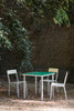 A beautiful garden on a sunny summer day. Minimalistic but colourful outdoor furniture in aluminium. Green table top. Three modern chairs around the table. Brick wall and leafy trees in the background.