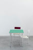 Minimalistic and colourful dining room furniture. Small table with green table top. Two chairs. Concrete floor. White wall.