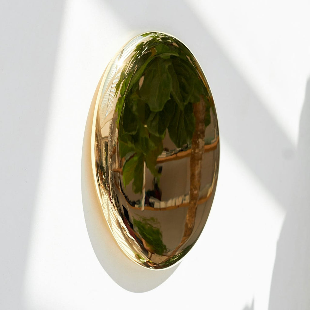 Kringlóttur og skúlptúrískur, kringlóttur spegill úr fægðu bronsi eftir Michael Anastassiades. Hvítur veggur.