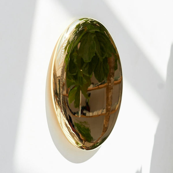 Kringlóttur og skúlptúrískur, kringlóttur spegill úr fægðu bronsi eftir Michael Anastassiades. Hvítur veggur.