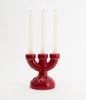 A ultra cute, three spoke candlebra in the colour burgundy with three white candles. The candles are lit. The background is white.