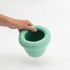 A hand holds up a tiny little ceramic planter. The planters colour is pale green. The planter is empty. White background.