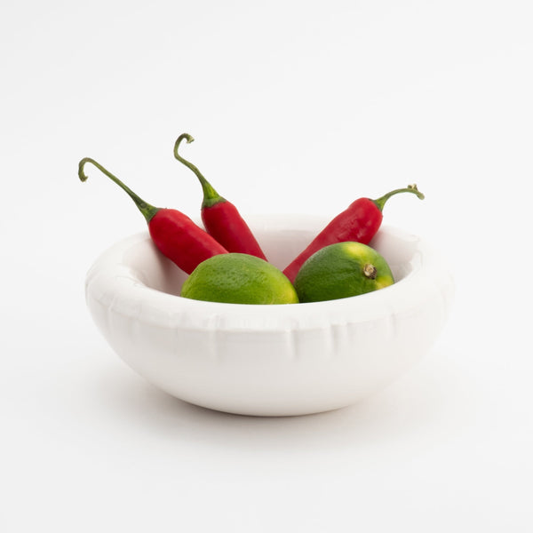 A white ceramic bowl with soft shapes. Red peppers and limes are in the bowl. White background.
