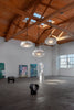 Three, large oval shaped pendants hanging next to eachother in various heights. Interior shot. Large and bright gallery space with white walls and high wooden ceilings. Light grey concrete floor. Some sculptures on the floor and some paintings in the background next to the wall.