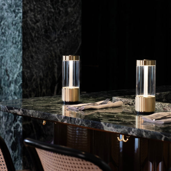 Tveir glæsilegir borðlampar úr brassi standa hlið við hlið á dökkri marmaraborðplötu. Kveikt er á lömpunum.