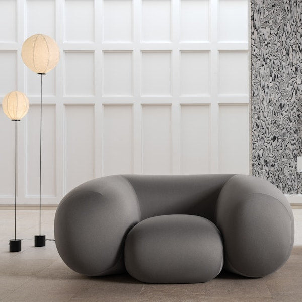 Fallegur og þægilegur, grár hægindastóll í mjúkum formum. Hvítur veggur í bakgrunni. Beige flísar á gólfi. Tveir gólfampar við hlið stólsins.