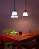 Two similar looking pendant lights side by side above a red, square dining table. Both lights are switched on. Wall in the background is in burgundy colour.