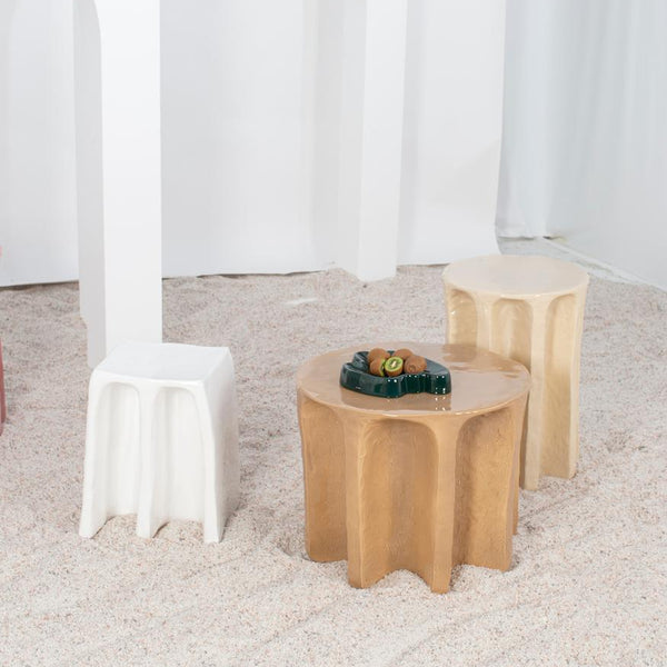 Skúlptúrísk húsgögn úr keramík. Hvítur kollur ásamt tveimur hliðarborðum. Hið fyrra er ljós brúnt en hið seinna er beige litað. Sandur á gólfi. Hvítur veggur í bakgrunni.