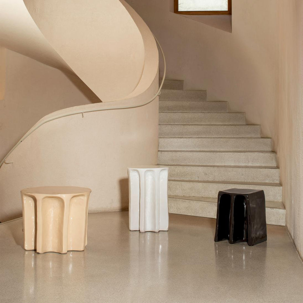 Tvö skúlptúrísk keramík hliðarborð ásamt einum skúlptúríkum kolli standa hlið við hlið. Fyrra borðið er beige litað, hið seinna hvítt og kollurinn er svartur. Stór stigi í bakgrunni.