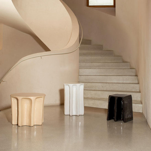 Tvö skúlptúrísk keramík hliðarborð ásamt einum skúlptúríkum kolli standa hlið við hlið. Fyrra borðið er beige litað, hið seinna hvítt og kollurinn er svartur. Stór stigi í bakgrunni.