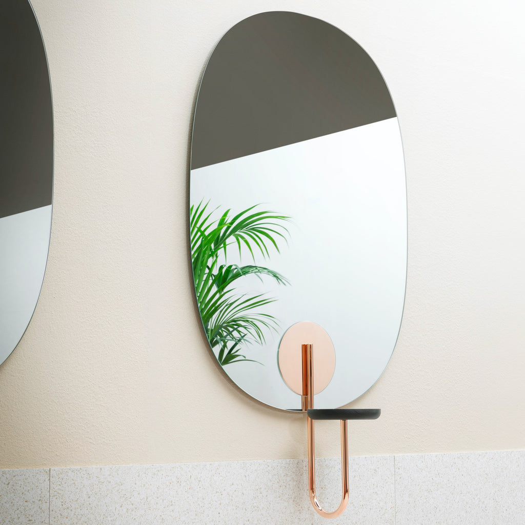 A big, gorgeous, oval shaped wall mirror with a build in flowerpot stand in copper. A pale coloured wall. A reflection of a flower in the mirror.