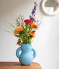 The cutest ceramic vase in mat, baby blue colour. Flowers in the vase. White wall in the background.