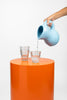 A hand poures water from a baby blue ceramic water jug into a clear glass. The glas sits on a orange plinth.