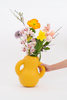 A hand reaches for a mat, yellow and curvy ceramic vase. White background. Some flowers in the vase.