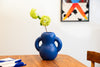 A curvy, navy blue ceramic vase sits on top of a dining table. A flower in the vase. A white wall in the background.