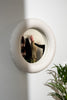 The cutest little wall mirror. The mirror is round and made from mat, white ceramics. The mirror hangs on a white wall. A reflection of a coat hanger.