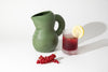 A cute ceramic water jug in the colour moss green on a white background. Next to the jug are some red berries and a glass with berry juice with a slice of lemon.