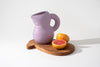 A cute, purple ceramic jug sits on top of a wooden cutting board next to a red grape. White background.