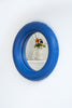 A blue, round, wall mirror hangs on a white wall. The mirror is made from mat, blue ceramics and has some cartoonish features. A reflection of a white ceramic jug on a shelf with flowers is in the mirror.