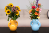 Two ceramic vases next to each other. The one on the left is yellow and glossy while the one on the right is mat, baby blue. Both of the vases have flowers in them.