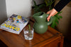 A mat, moss green ceramic jug sits on top of a side table along with magazines and a glass of water. A hand reaches for the jug.