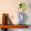 White vase with two handles holds a single stem of white hydrangea, placed on a wooden shelf with books underneath.