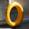 A cute, round mirror made from mat, yellow ceramic stands on the ground. A plexiglass wall in the background.