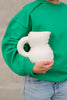 A woman wearing jeans and a green sweat shirt hold a cute jug made from mat, white ceramics.