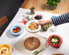 A collection of five, round ceramic bowls on a table. The baby blue bowl has tomatoes. The pink bowl has olives. The white bowl has crackers. The red bowl has yoghurt salat. The yellow bowl has hummus.