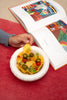 A round, white ceramic bowl on a table next to a open artbook. A hand reaches for some tortilla chips from the bowl.