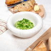 A round, white ceramic bowl on a table. Pea salat in the bowl. A loaf of bread next to the bowl.
