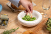 A round, white ceramic bowl on a table. Pea salat in the bowl. Bread, sardines and a glass of water on the table. A hand reaches for the bowl.