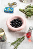 A rounded, pale pink ceramic bowl on a table. Black olives in the bowl.