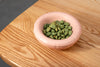 A round, pale pink ceramic bowl on a table filled with green wasabi nuts.